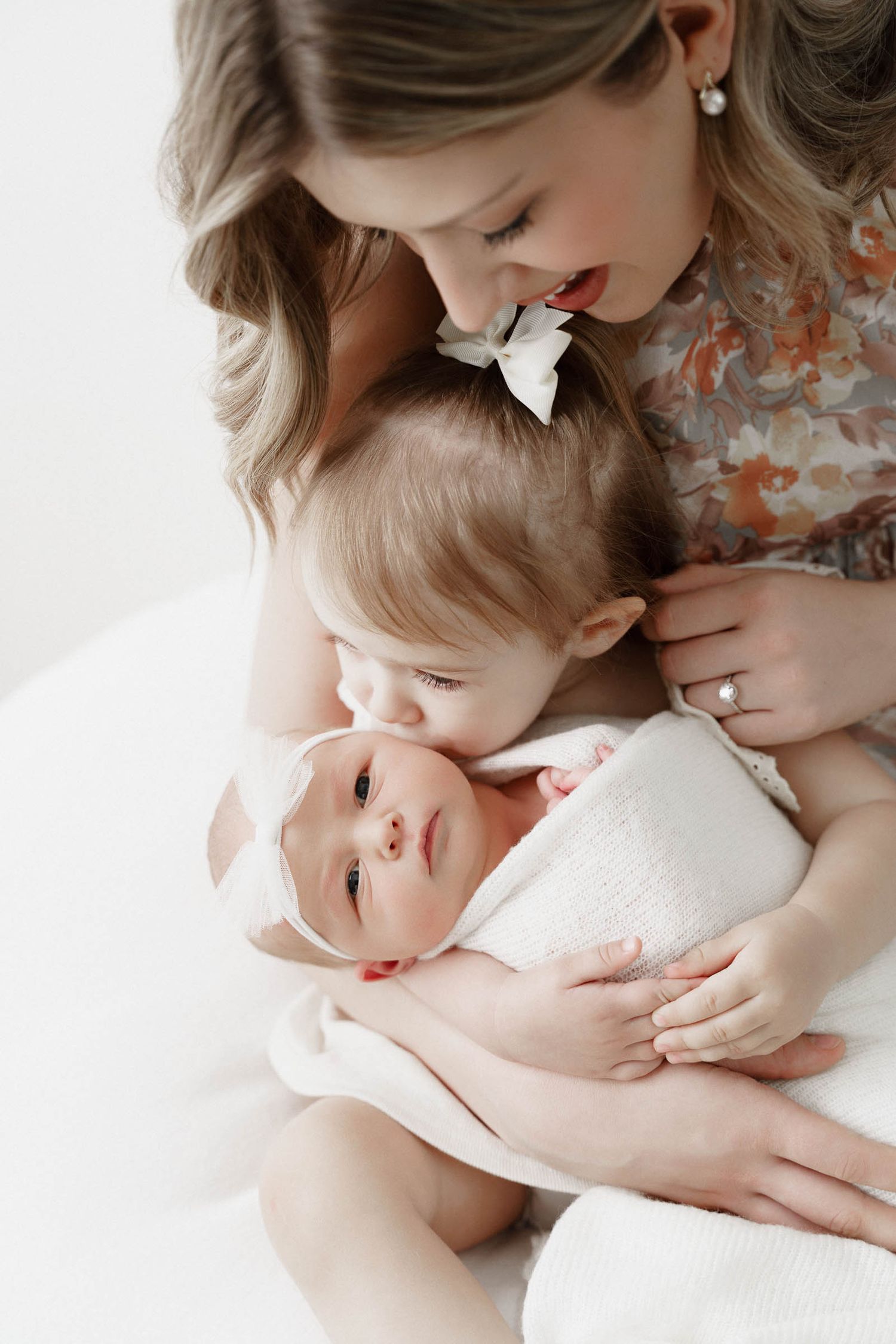 Mother in floral dress tenderly holding and gazing at baby in white against light background.