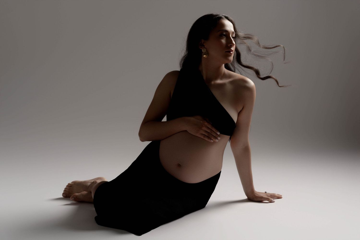 Artistic maternity photos showing pregnant figure in black dress reclining on light floor.