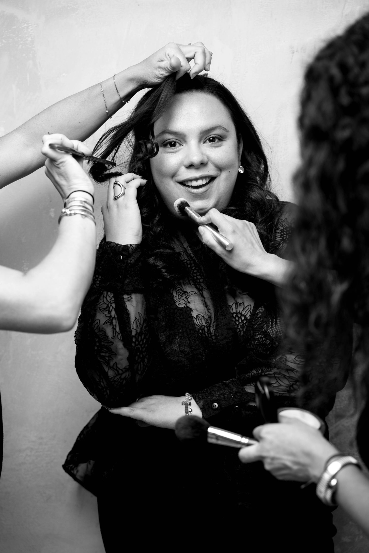 Black and white photo of a smiling woman getting hair and makeup done by two stylists backstage.