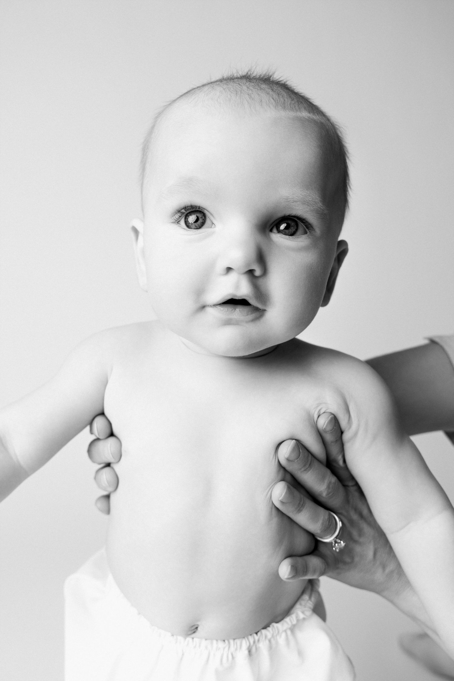 Black and white photo of an alert baby in a diaper being held up by adult hands wearing a ring.