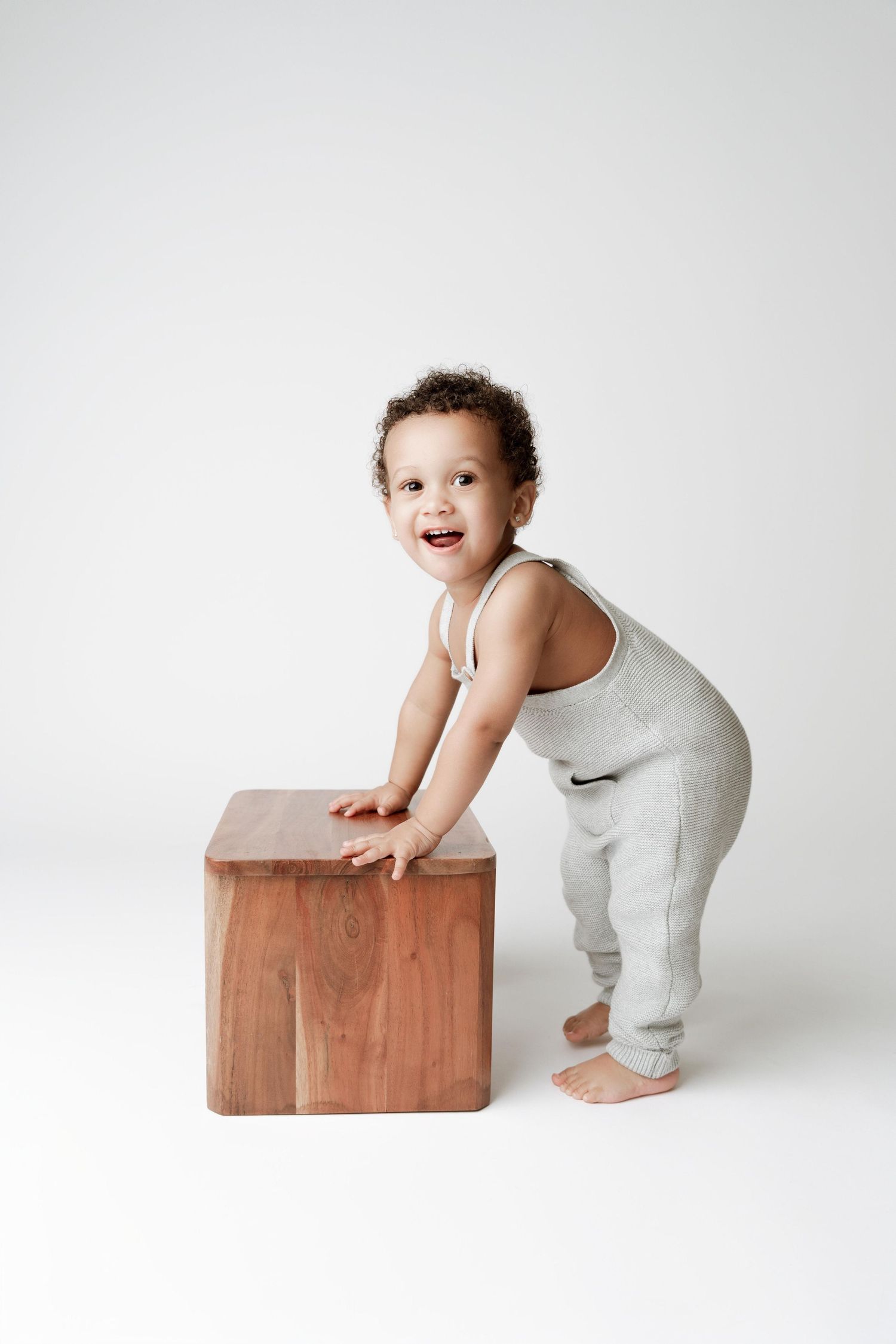 Smiling toddler in gray outfit standing barefoot next to a wooden cube on a white background.