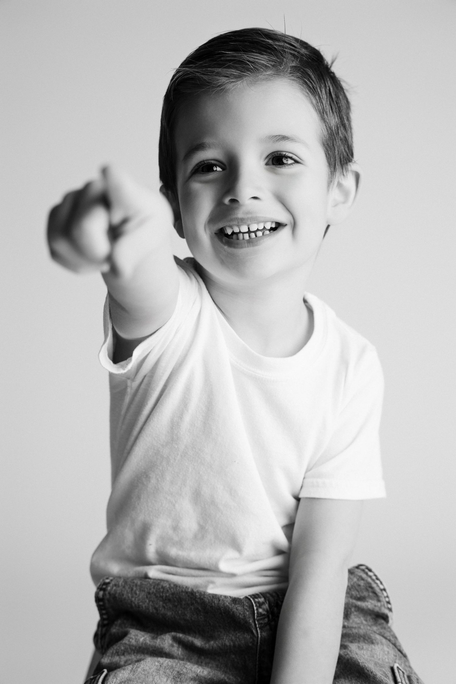 Black and white photo of a happy young boy in white t-shirt pointing at camera and smiling.
