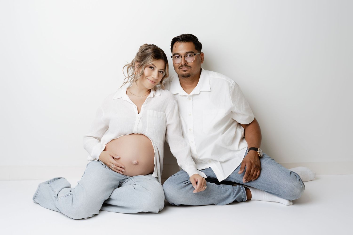 Expecting couple in matching white shirts and jeans sit together against white background during maternity photoshoot.