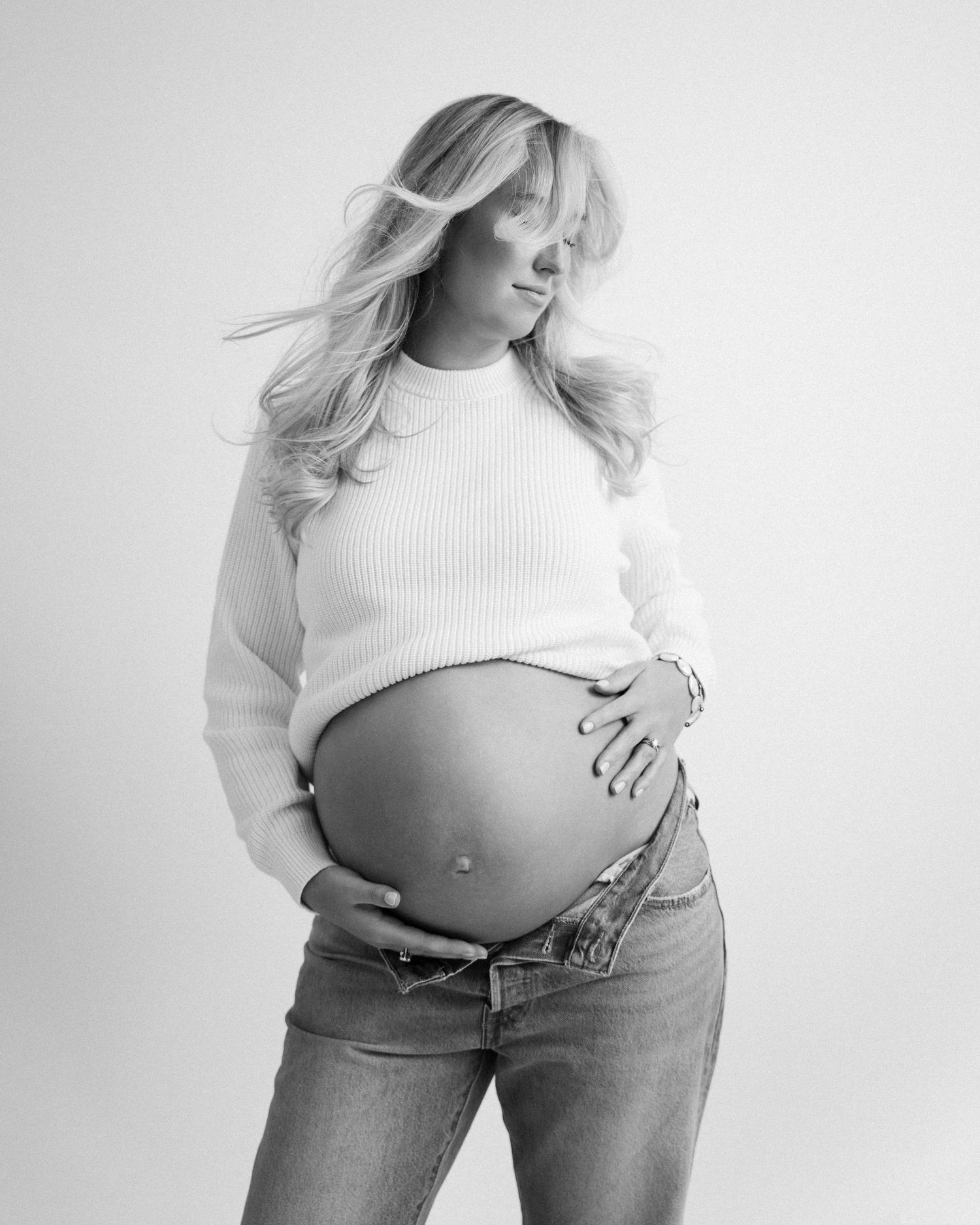 Black and white maternity photo showing pregnant woman in white sweater and jeans holding her bare belly.