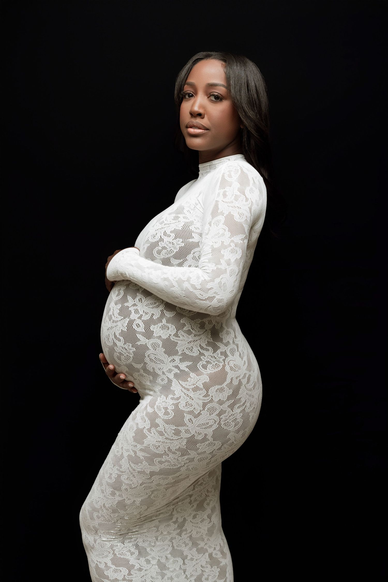 Dramatic maternity portraits with woman in white lace gown against dark background.