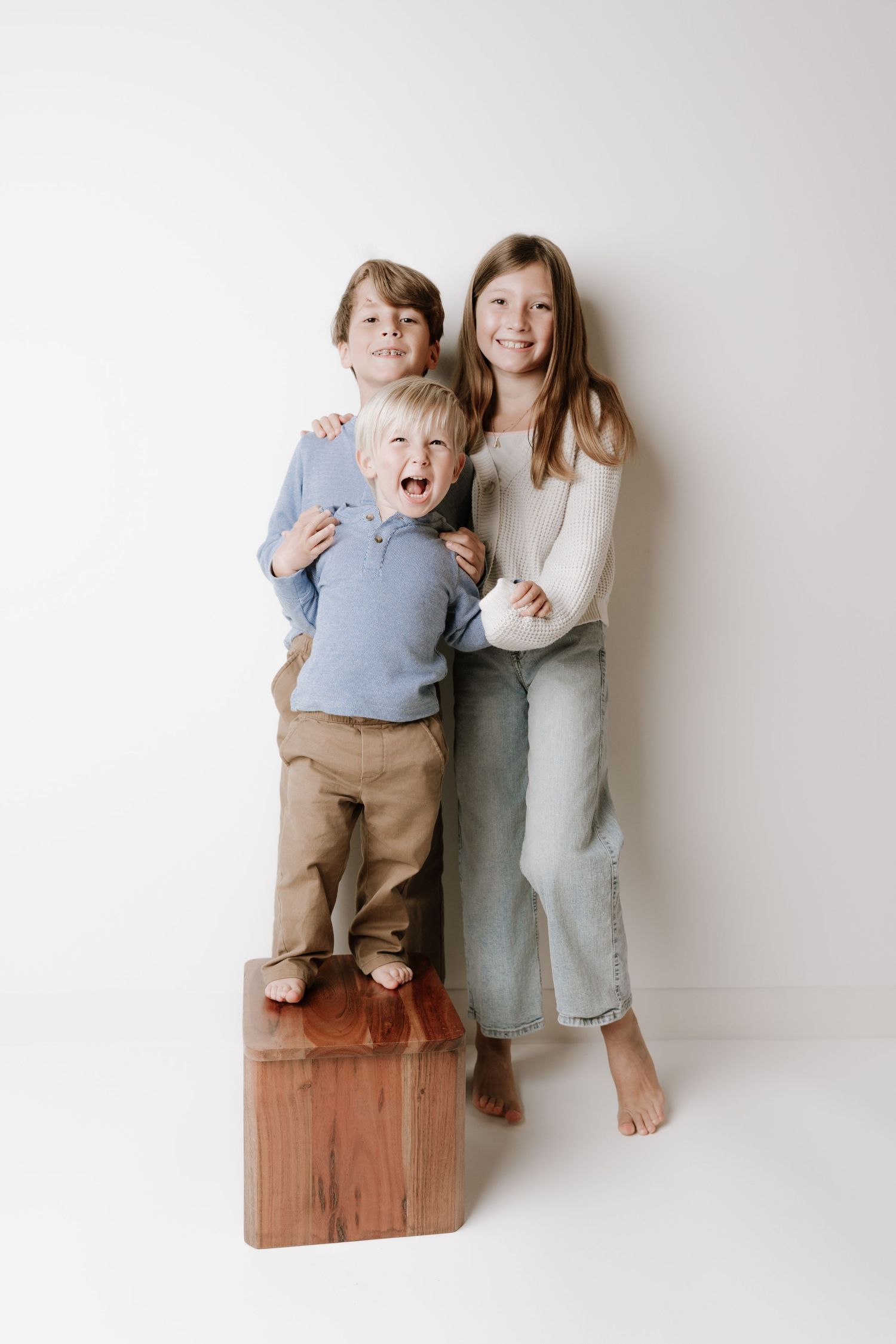 Three barefoot children pose together against a white background, two older kids hugging a laughing toddler on a wooden block.
