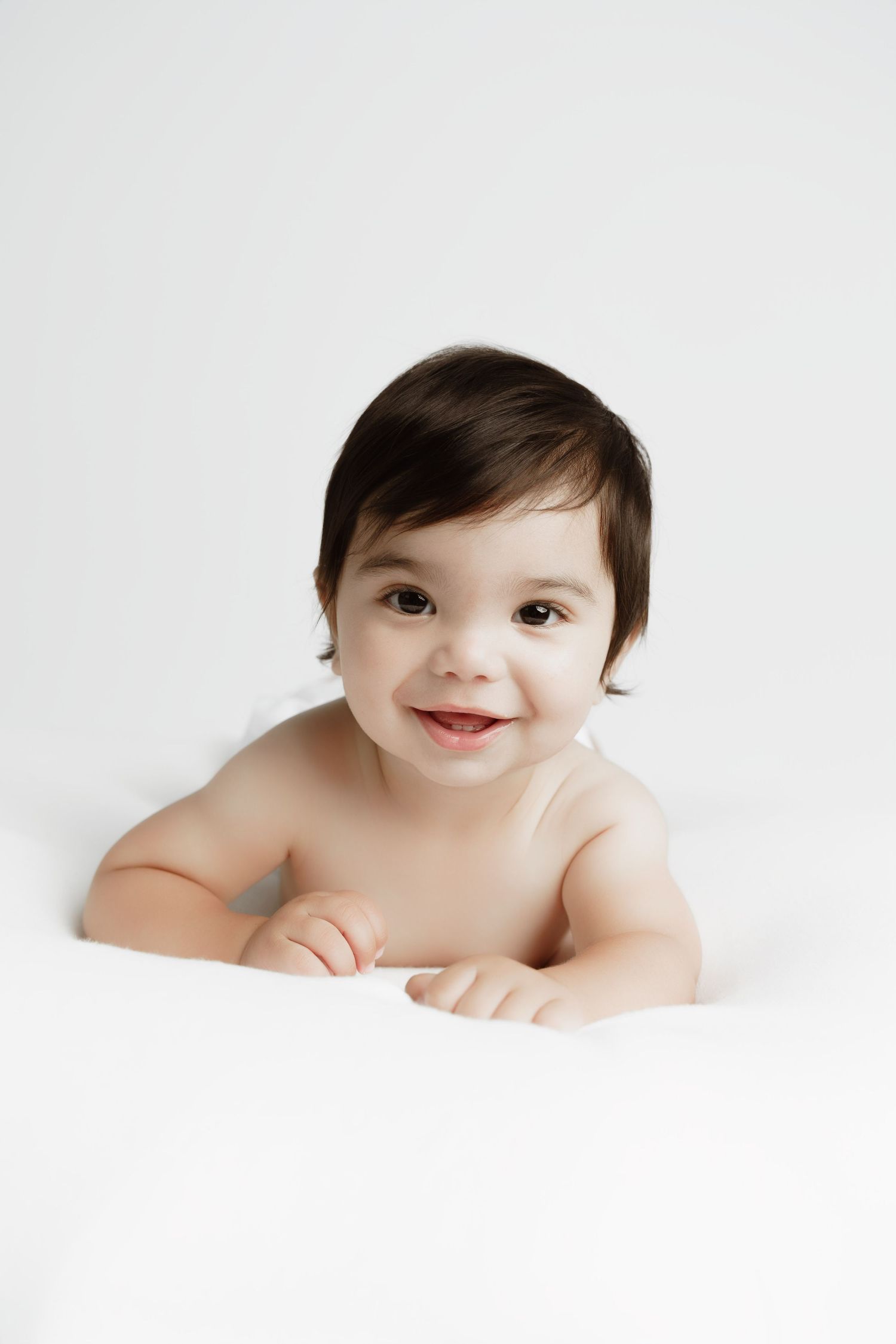 Smiling baby with dark hair lying on white surface, looking at camera with bright eyes and cheerful expression.