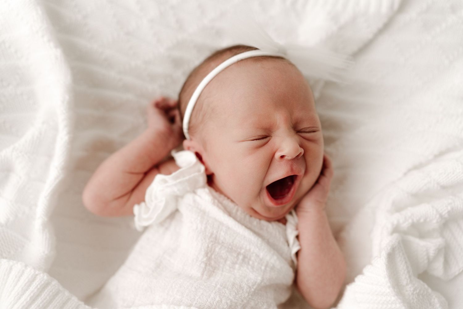 Newborn baby girl in white outfit and headband yawning while lying on a soft white blanket.