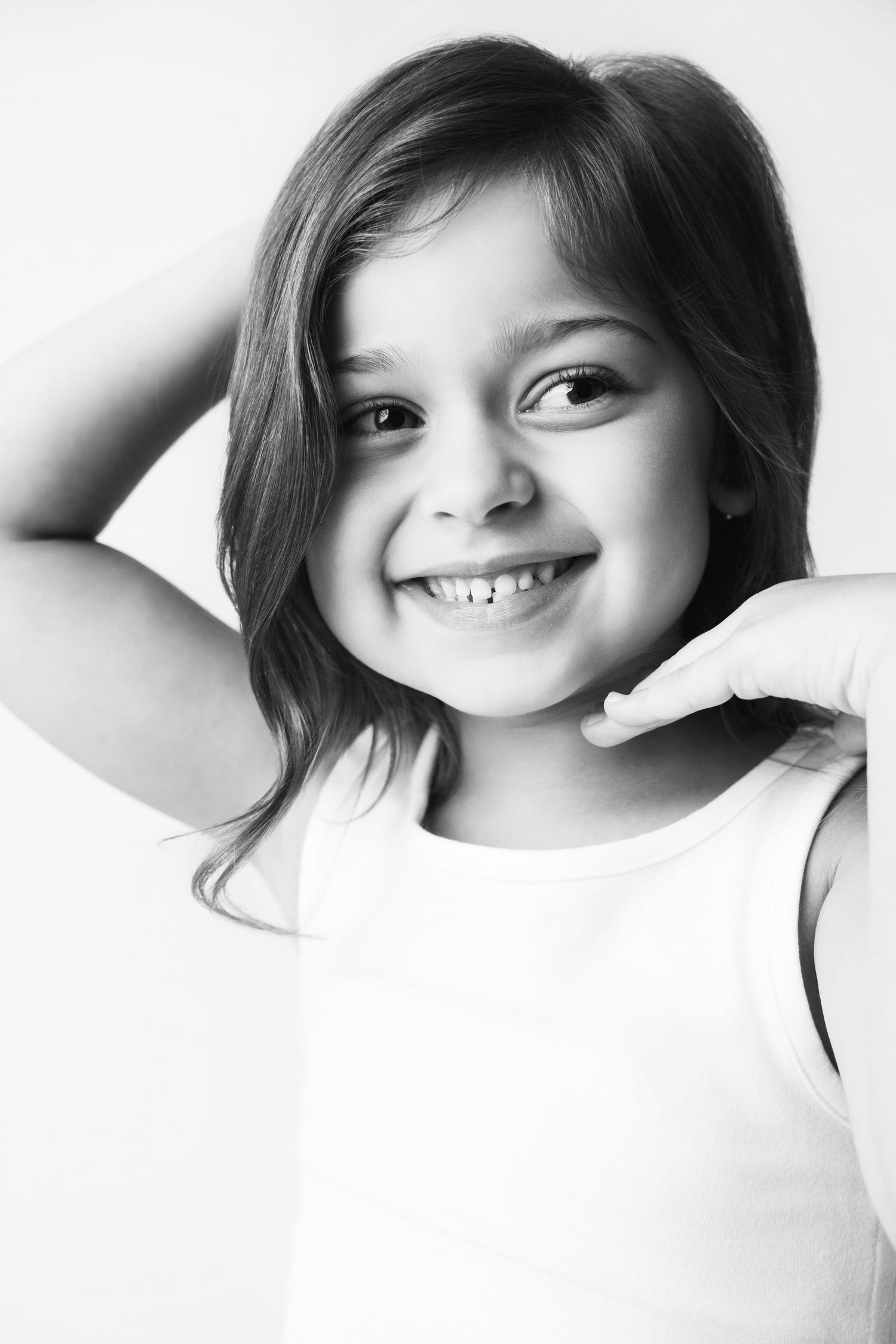 Black and white portrait of a smiling young girl in a white tank top with hand raised near face.