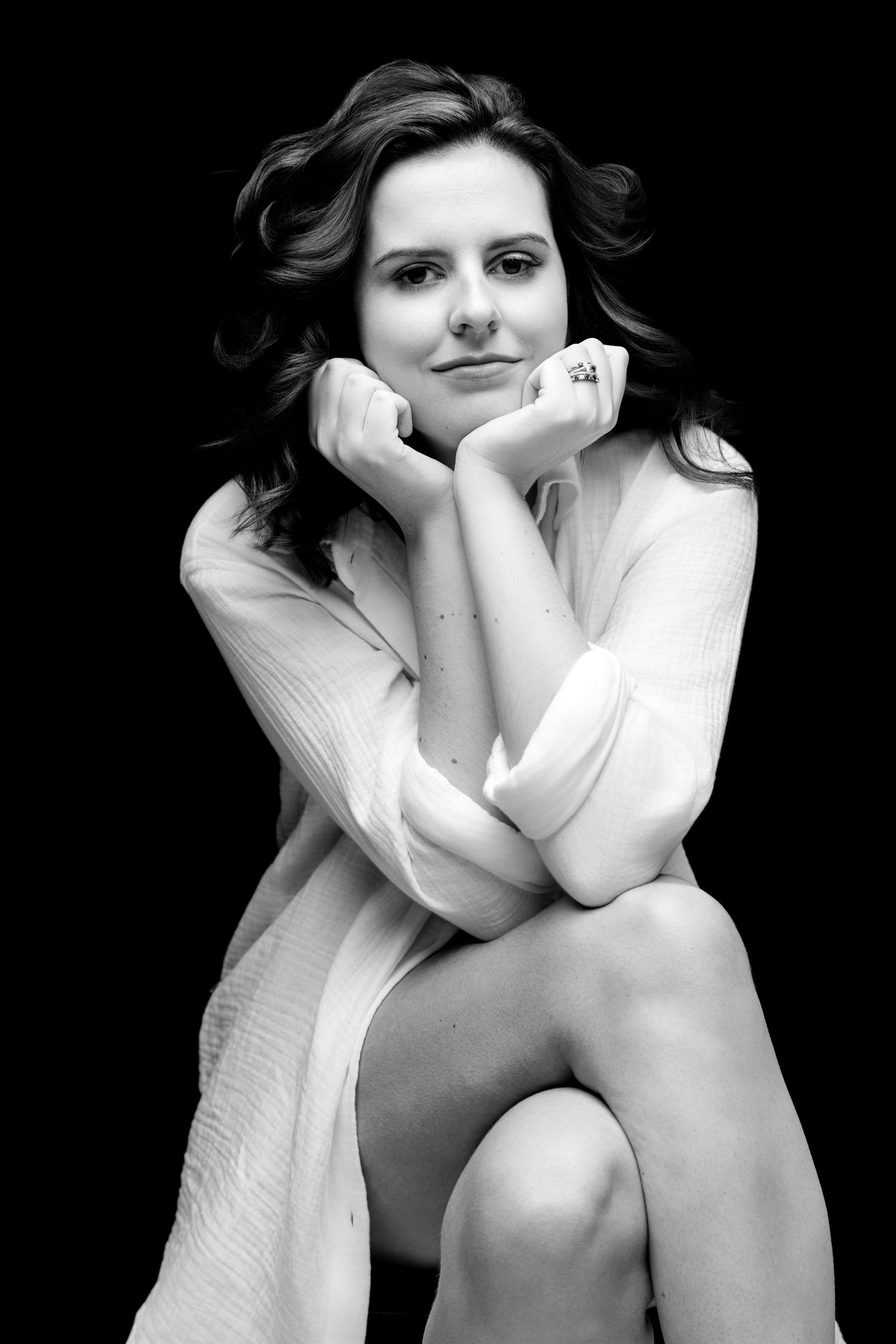 Black and white portrait of a young woman in a knit sweater, sitting with hands under chin, smiling softly.