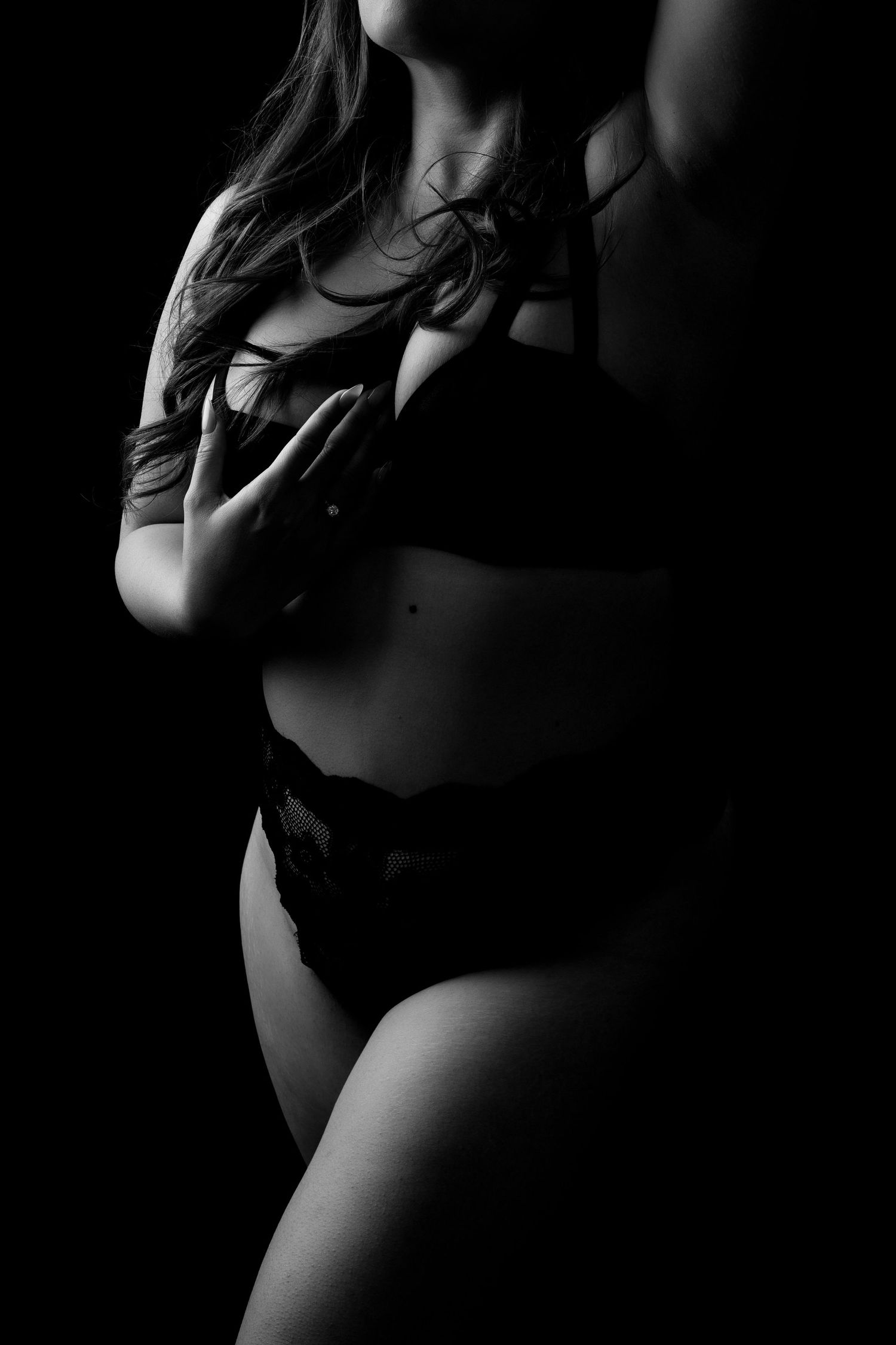 Black and white artistic photo of a pregnant woman in lingerie posing gracefully against a dark background.