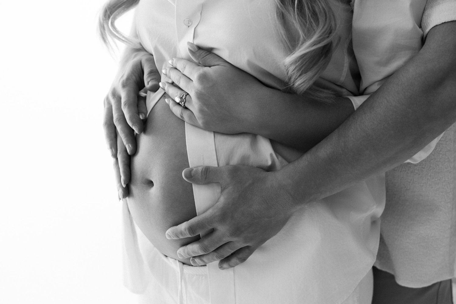 Black and white maternity photo of couple embracing, hands cradling pregnant belly.