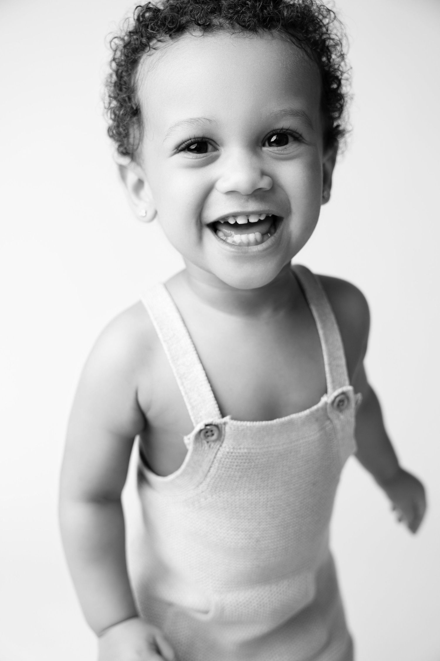 Black and white portrait of a happy toddler with curly hair wearing a knit romper, smiling brightly.