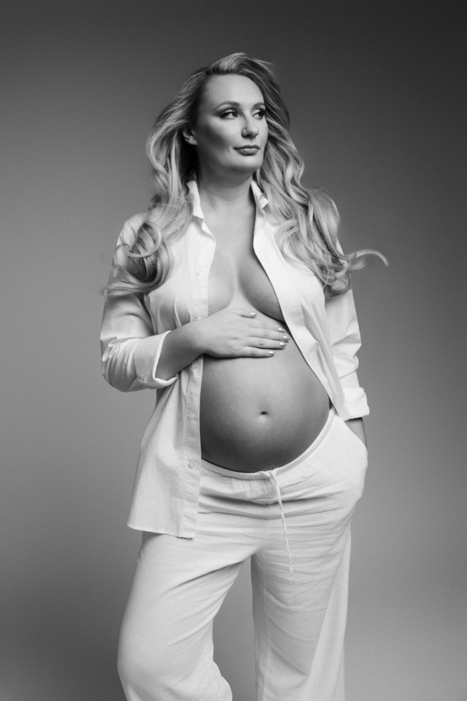 Black and white portrait of a pregnant woman in an open white shirt cradling her baby bump in a studio.