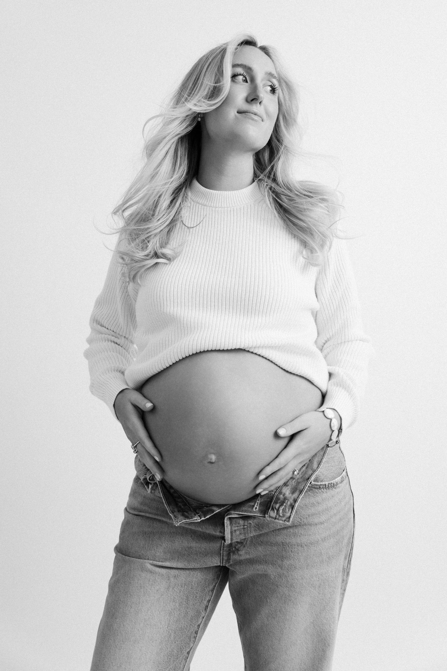 Black and white maternity photo of a woman in a white sweater and jeans showing her pregnant belly.