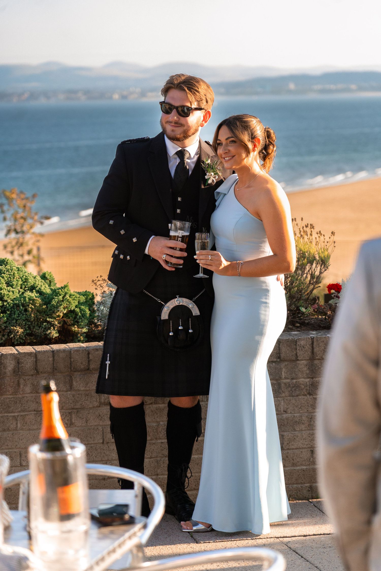 Couple in wedding attire toasting with drinks on a scenic oceanfront terrace.