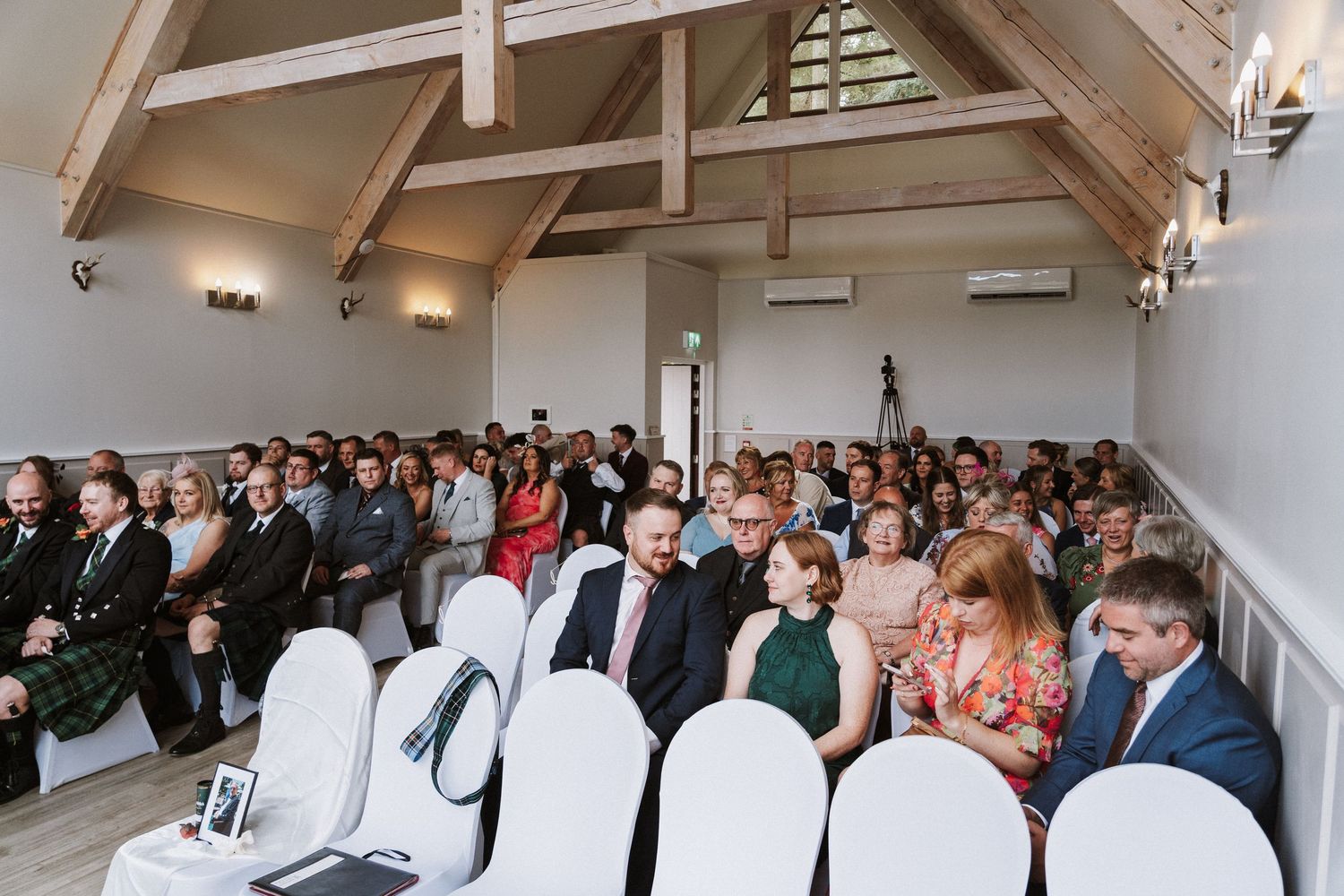 Wedding guests seated in rows during an indoor ceremony with white walls and warm lighting.