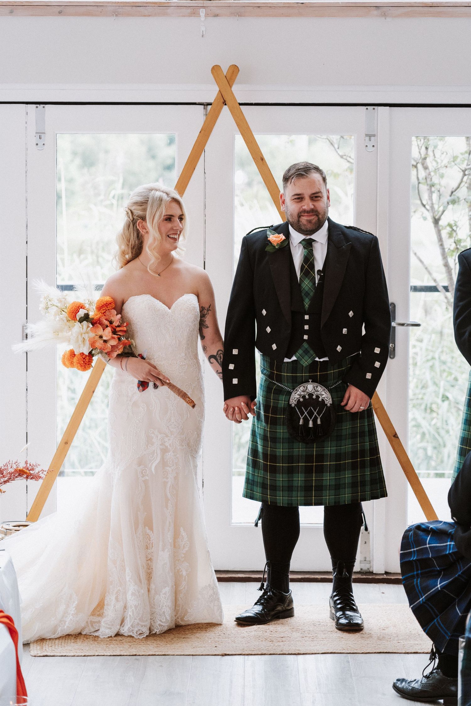 A wedding couple poses under a wooden arch, with the groom wearing a traditional Scottish kilt and the bride in a white gown.