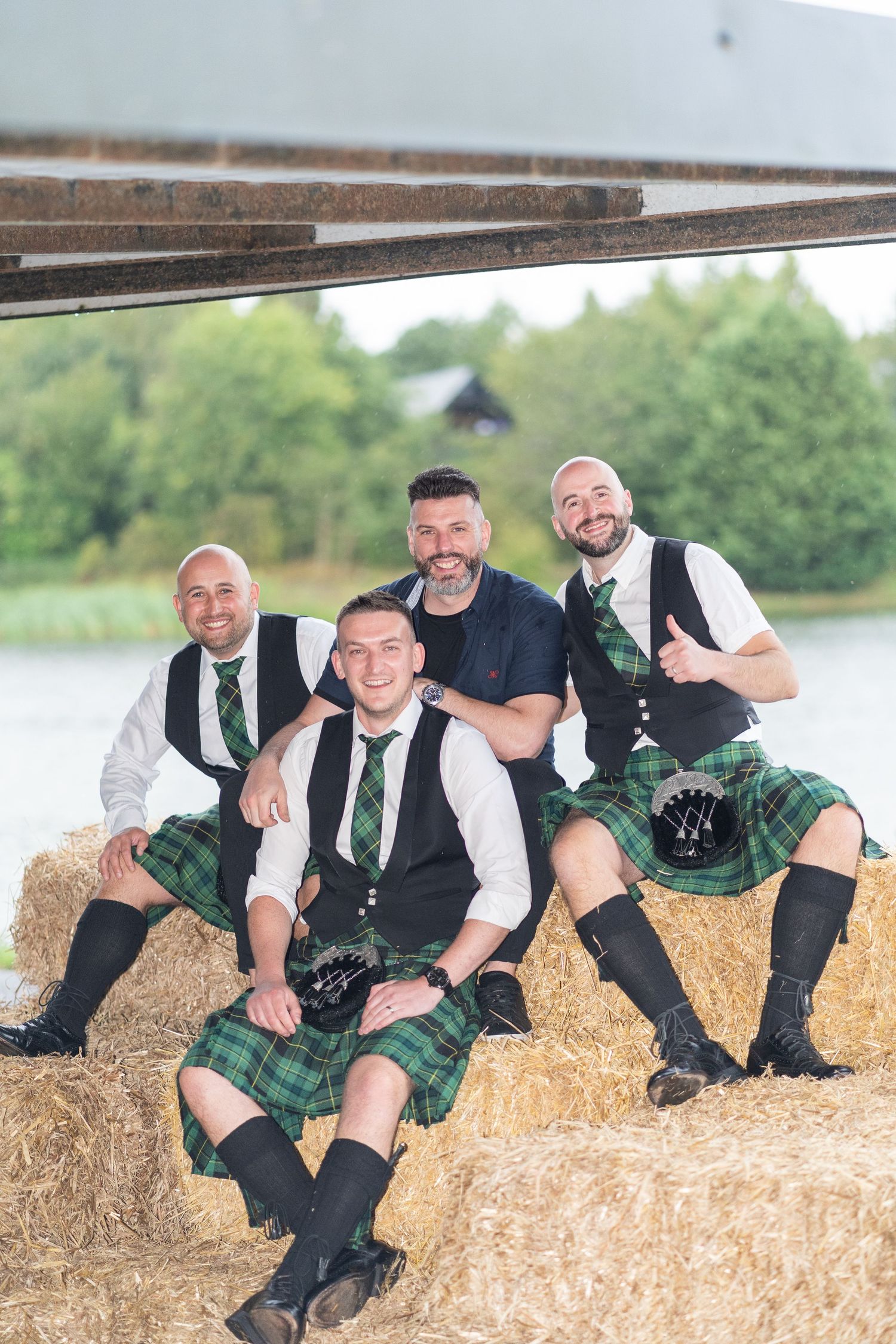 Groomsmen in kilts and vests lounging playfully on hay bales during outdoor wedding photo session.
