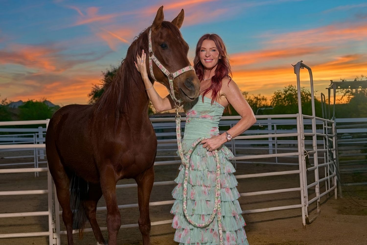 Young woman in ruffled mint gown poses with chestnut horse at sunset near metal fence.