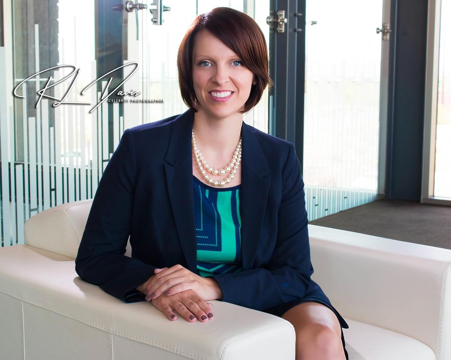 Professional portrait of woman in navy blazer and teal top seated in modern office setting. Photographer RL Davis