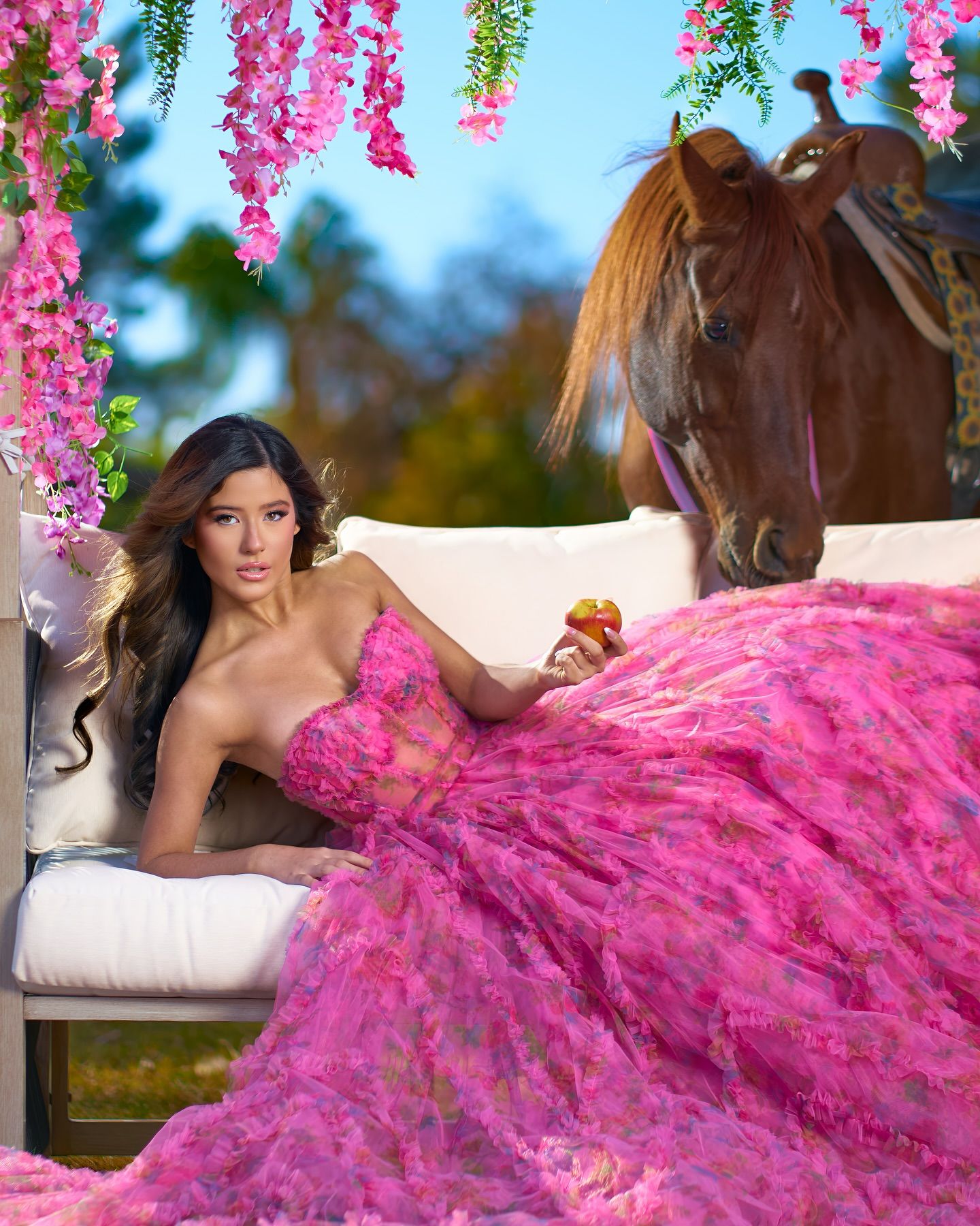 Woman in pink floral gown reclines on white settee beside a brown horse, surrounded by pink hanging flowers.