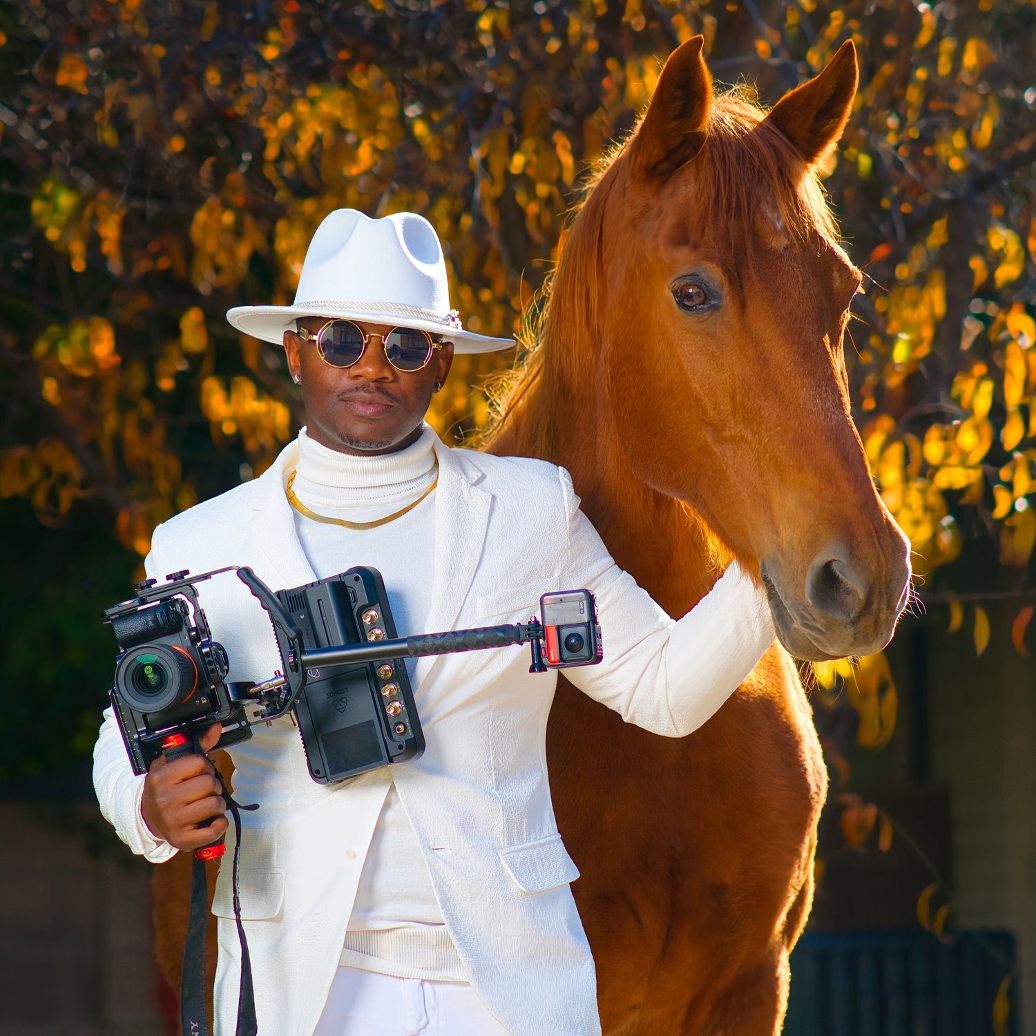 Professional photographer in white attire poses with camera equipment and a brown horse in autumn setting. Photographer RL Davis