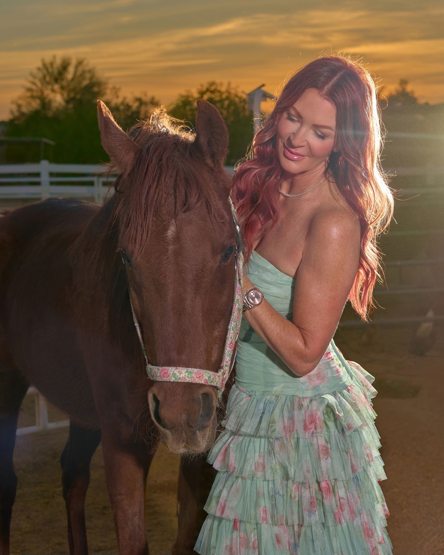 Woman in floral green dress poses with chestnut horse at golden sunset near ranch fence.