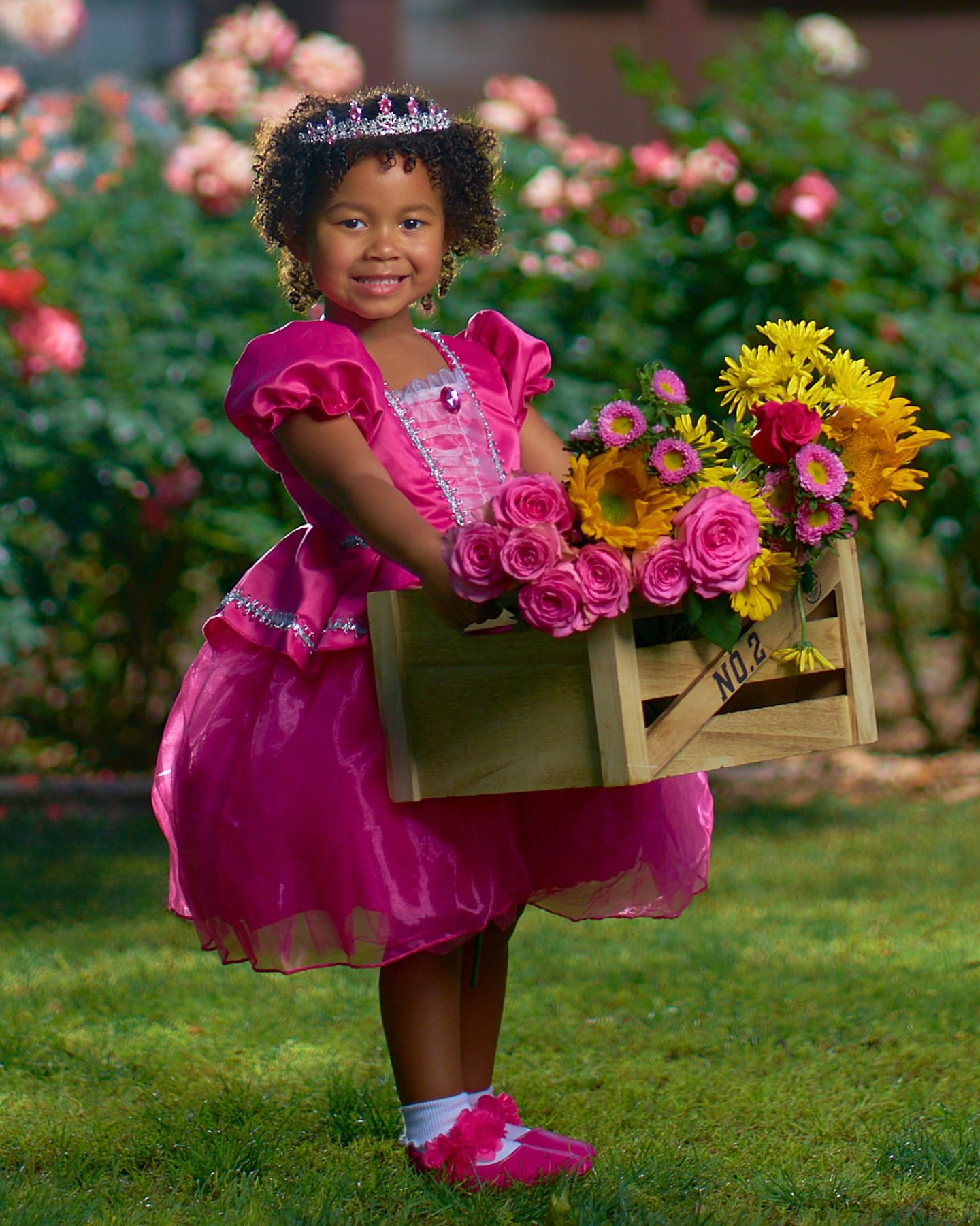 A child in a pink princess dress carries a wooden crate filled with colorful flowers in a garden. Photographer RL Davis