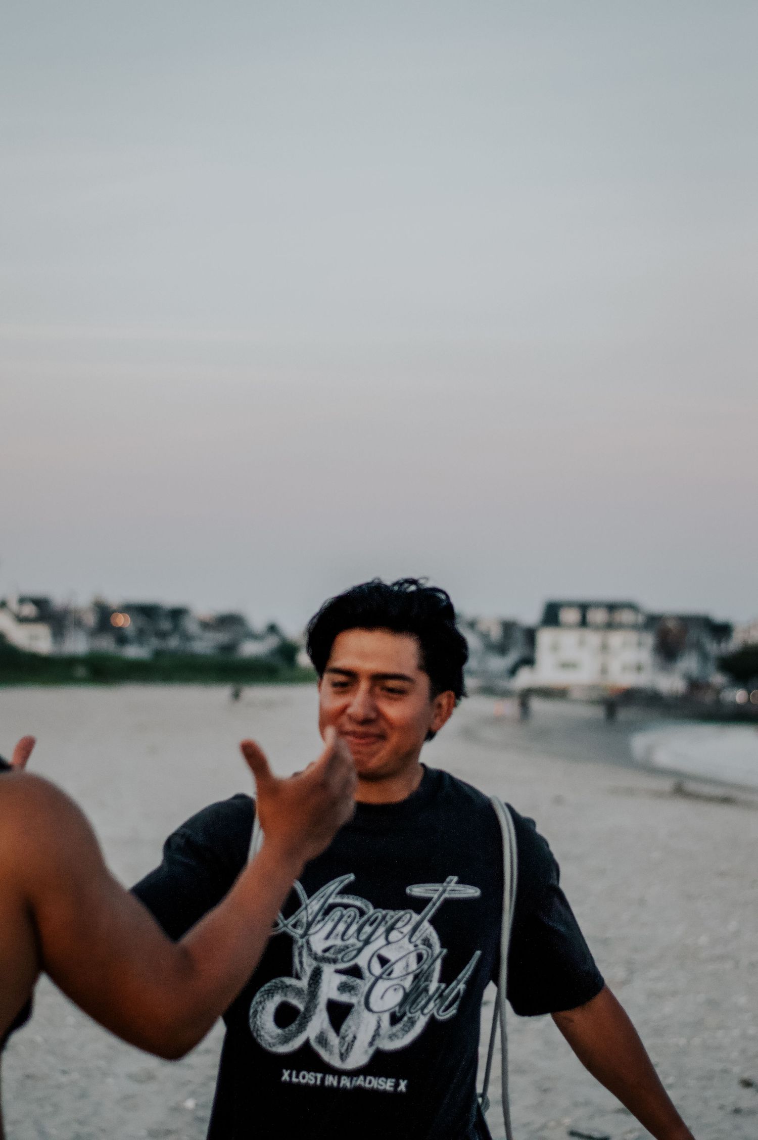 Young man in black graphic tee smiling on a beach at dusk, with coastal buildings in the background.