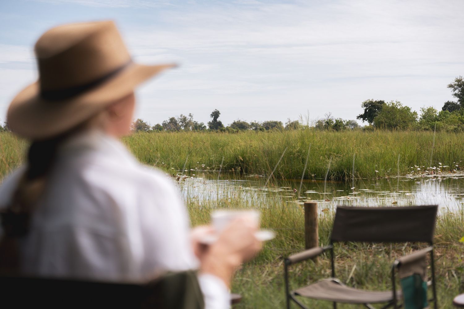 Person in hat sits by wetlands observing nature with binoculars.