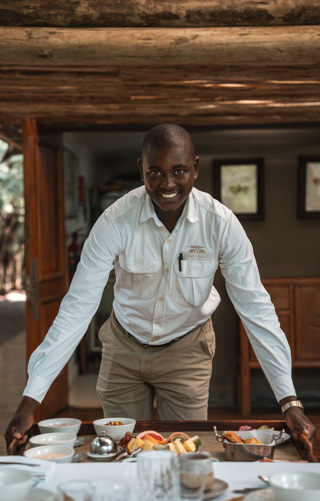 Staff member in white uniform shirt presents dining table set with fresh breakfast spread.