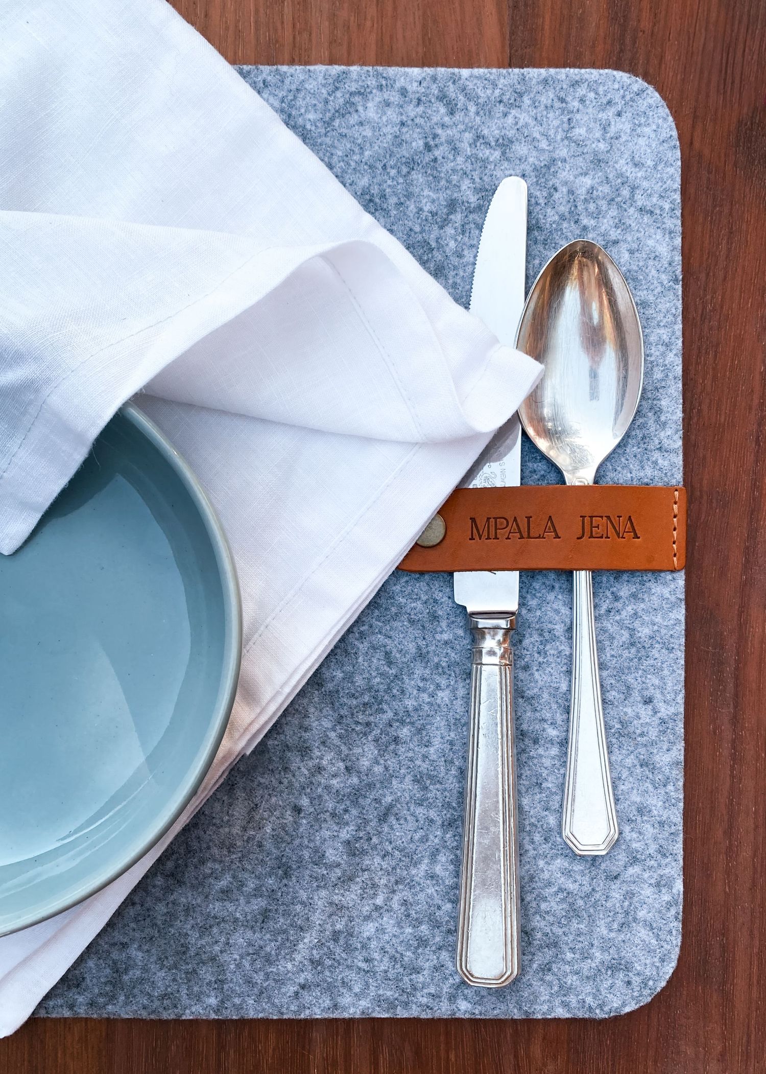 Place setting with blue plate, white napkin, and silverware on gray felt placemat with leather holder.