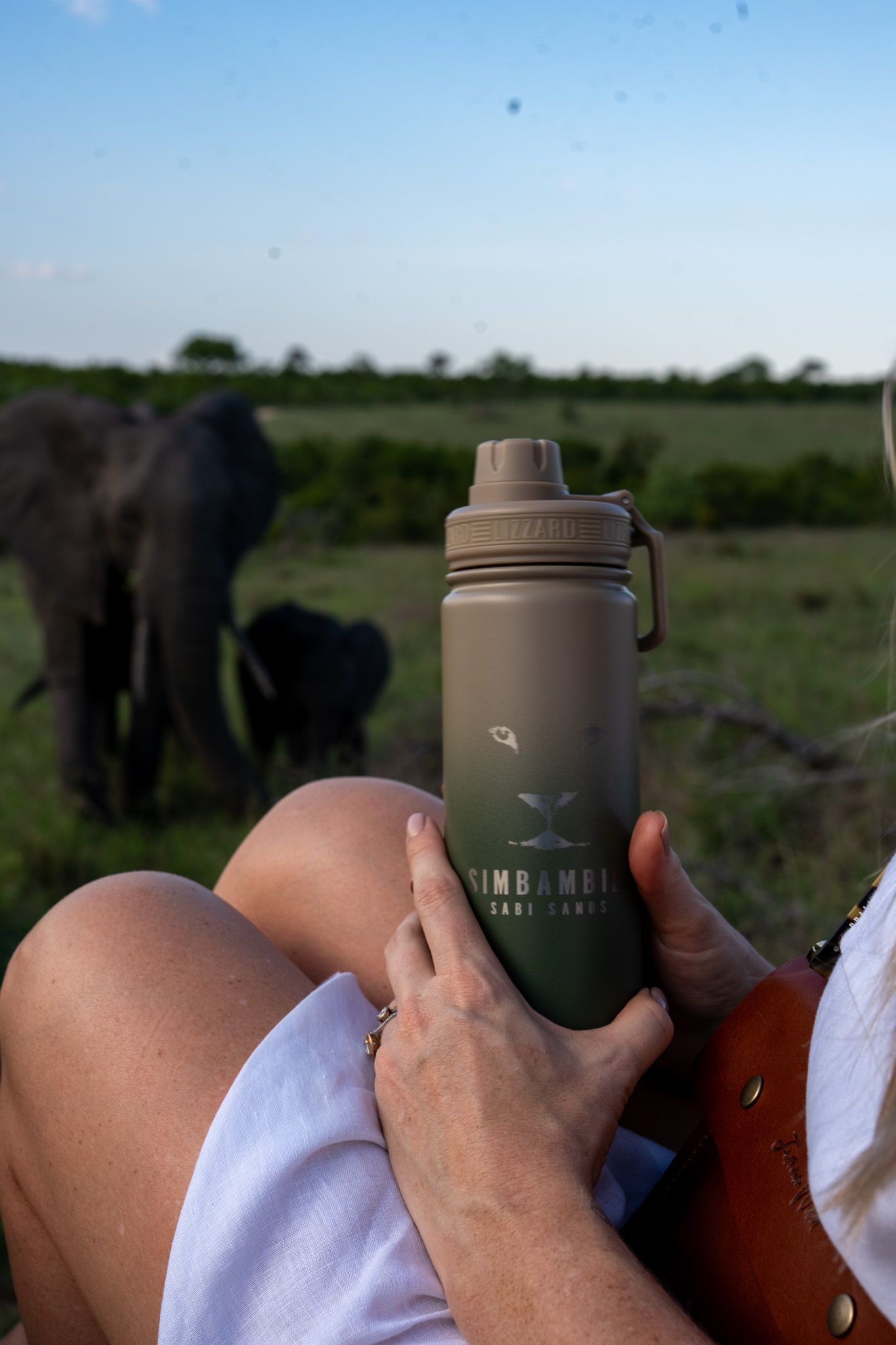 Metal water bottle held against safari landscape with elephant silhouette in background.