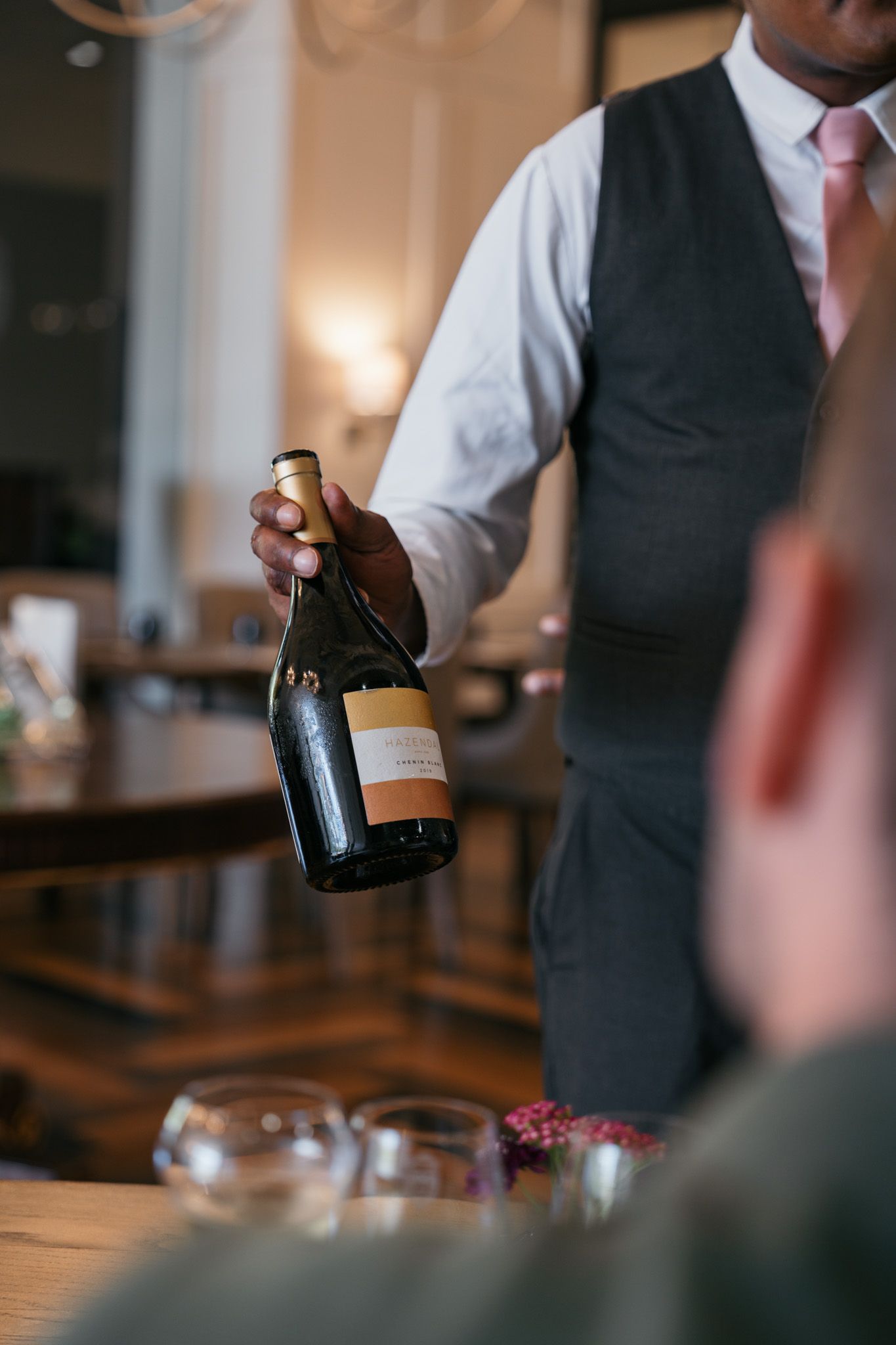 Server in formal attire holding a wine bottle in an upscale restaurant setting.