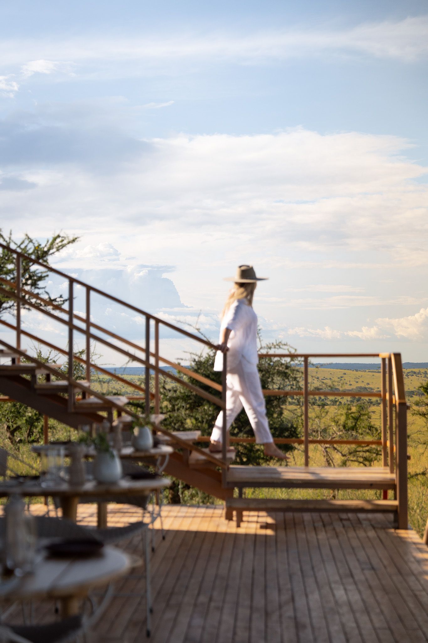 A white-clad figure ascends wooden stairs on an elevated deck overlooking a scenic natural landscape.