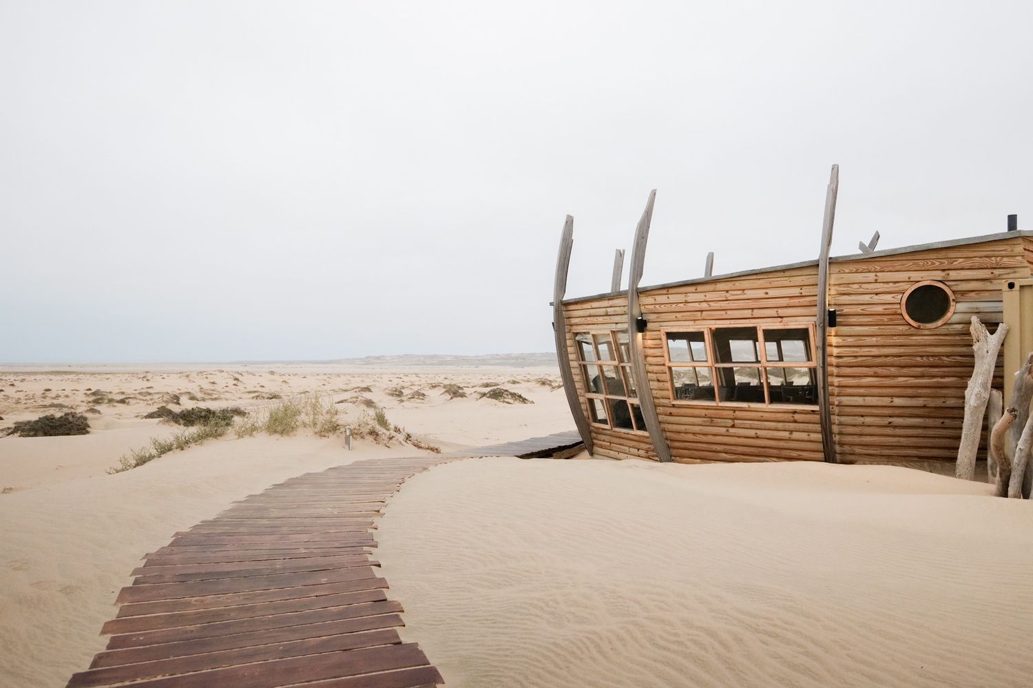 Weathered wooden beach house structure with boardwalk path through sandy desert landscape.