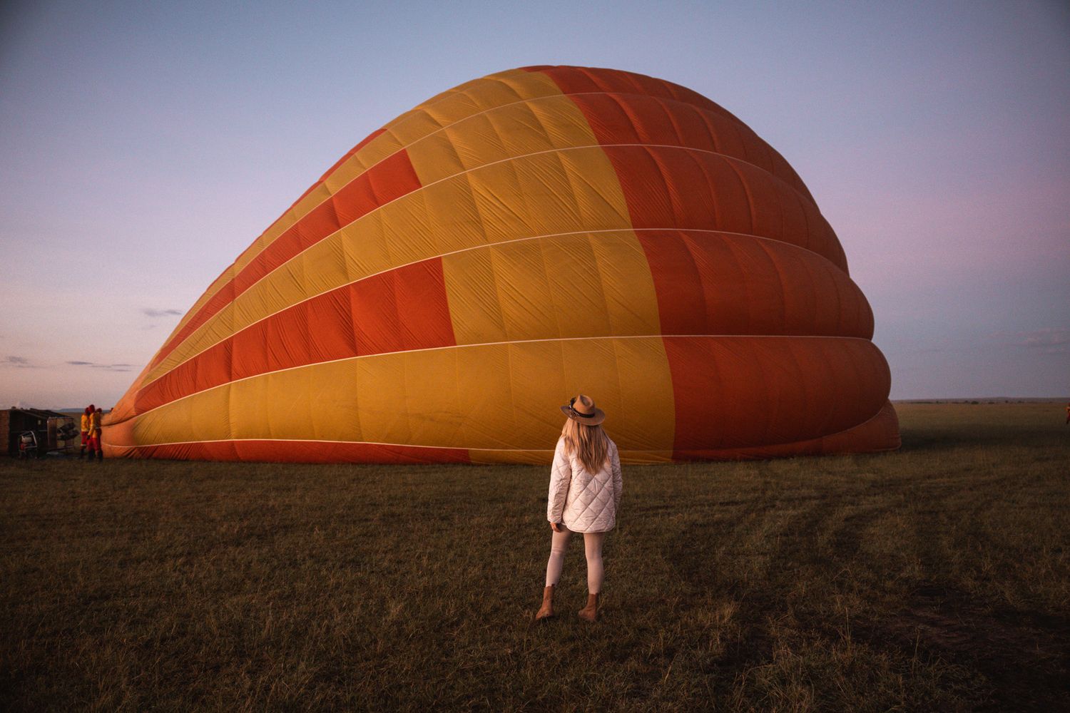 Large orange hot air balloon being inflated on grassy field at dusk.
