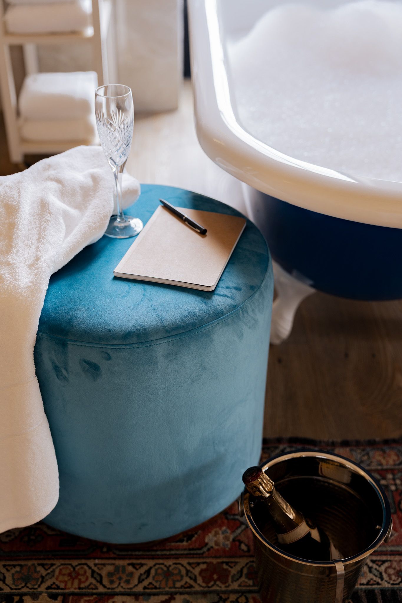 Luxury travel setup with champagne glass and notebook on turquoise velvet ottoman.