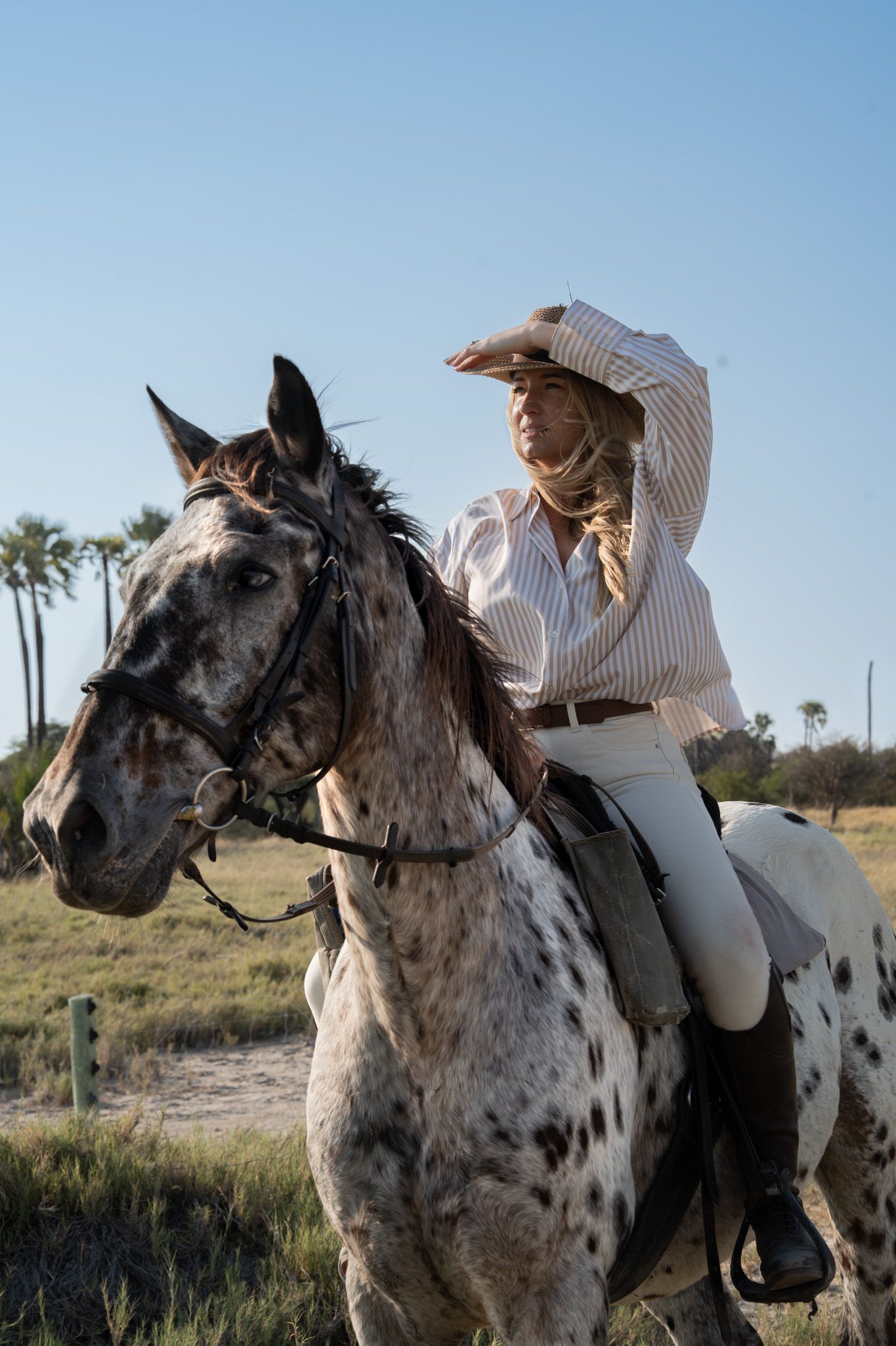 Rider atop spotted horse in white outfit against rural background with palm trees.