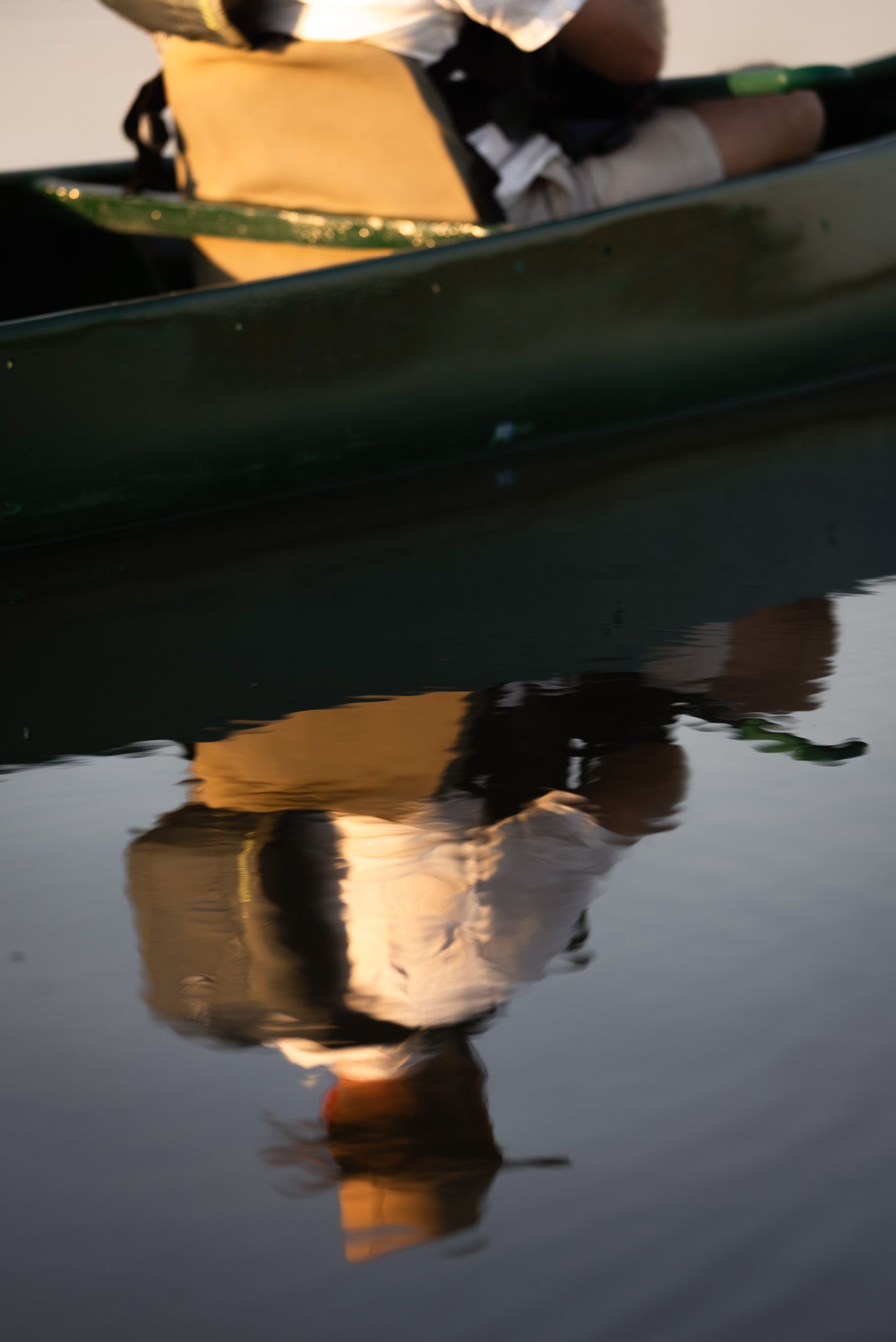 Reflection of canoe and paddler in calm water during sunset.