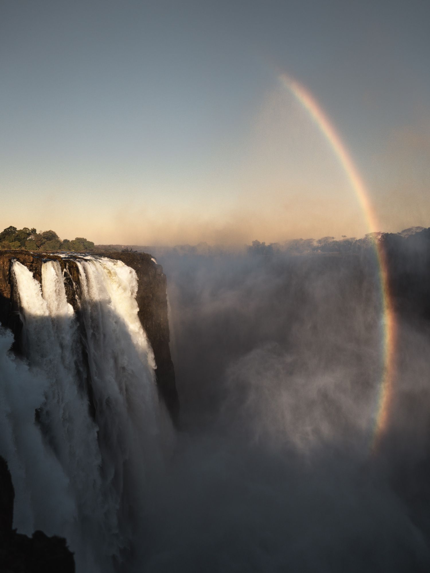 Dramatic rainbow arcs over Victoria Falls with misty spray rising from the massive waterfall.