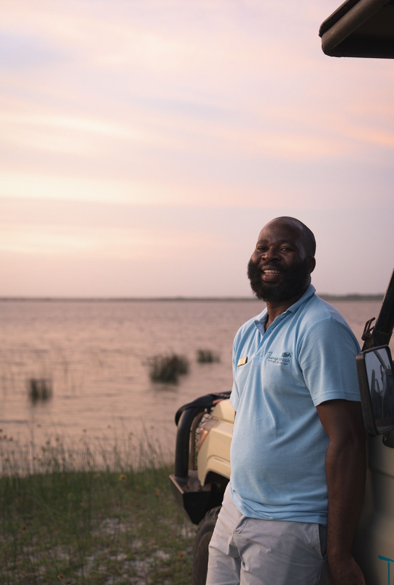 Person in light blue polo shirt standing by vehicle during scenic sunset over savanna landscape.