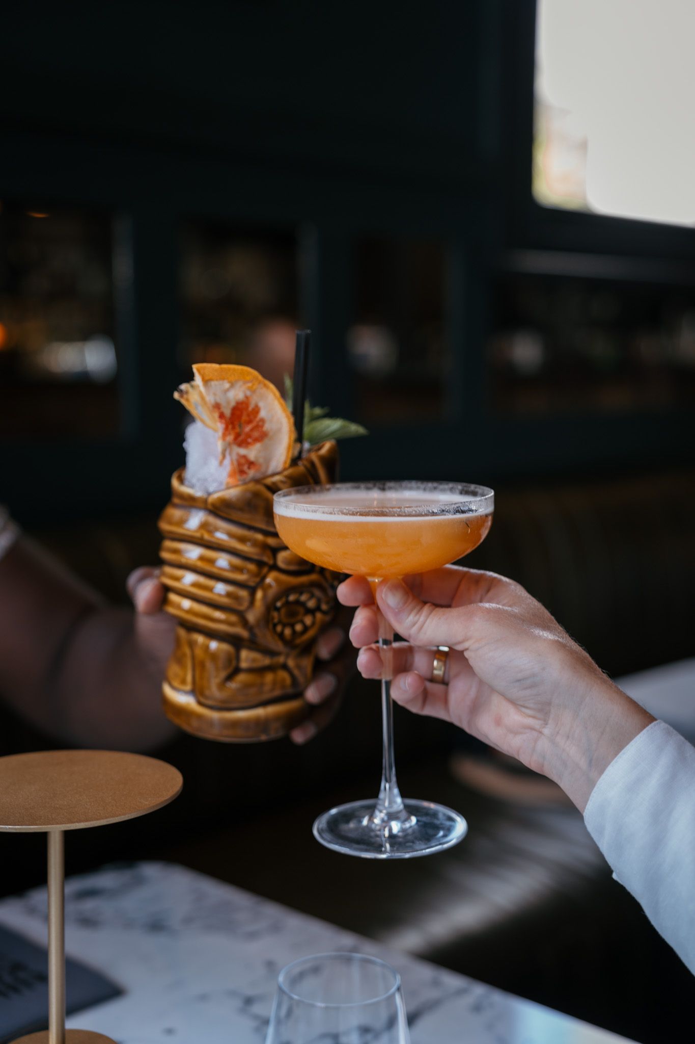 Tropical cocktail served in coupe glass next to decorative tiki mug.