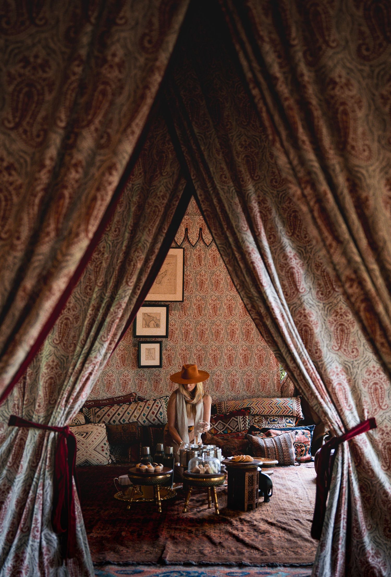 Woman in orange hat sits inside ornate Moroccan-style tent with paisley drapes, plush sofas, and decorative rugs.