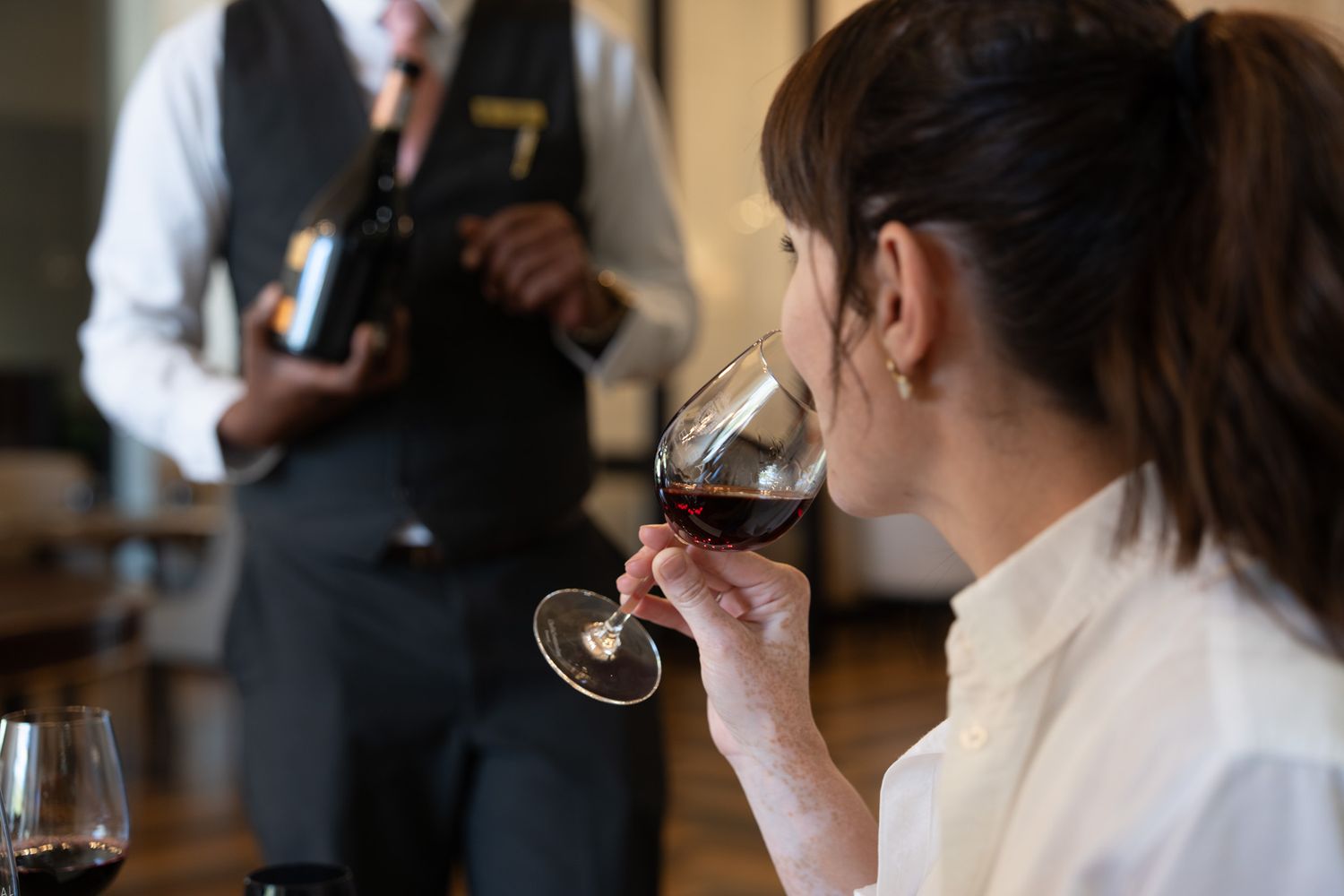 Guest tasting wine while sommelier presents bottle in upscale restaurant setting.