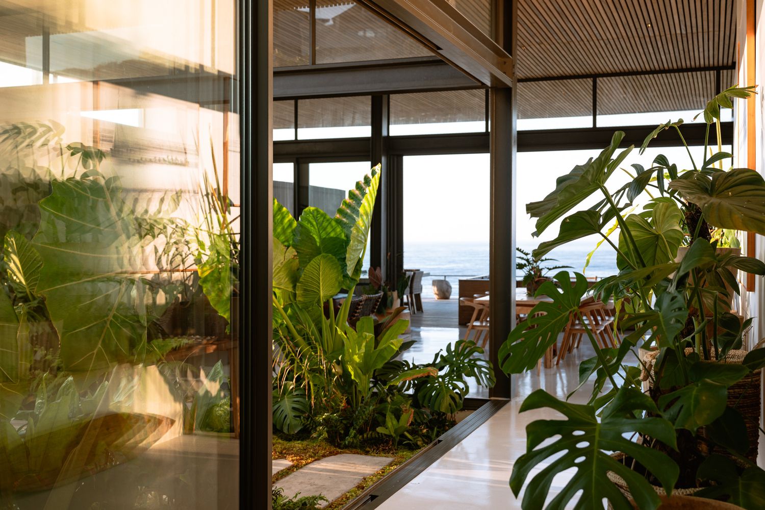 Interior walkway with lush indoor plants framing an ocean view through glass windows.