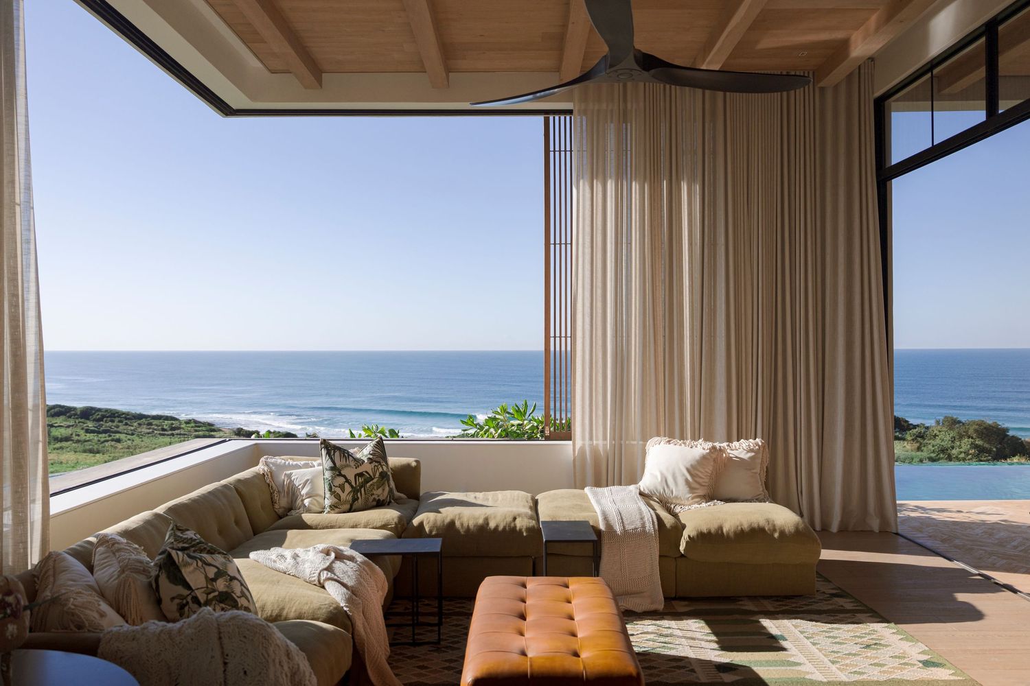 Luxury living room with ocean view, beige sofas, sheer curtains, and a leather ottoman on a sunny day.