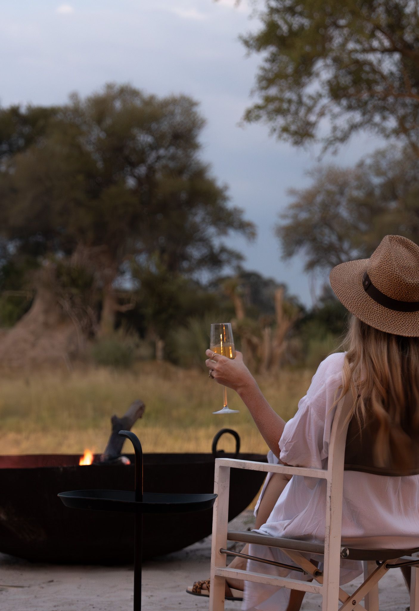 A person in a brown hat relaxes with a glass of champagne by an outdoor fire pit at dusk in a nature setting.