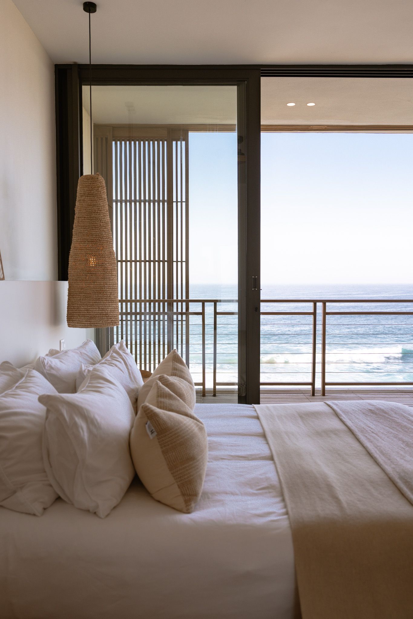 Serene oceanfront bedroom with white bedding and natural pendant light overlooking waves through floor-to-ceiling windows.